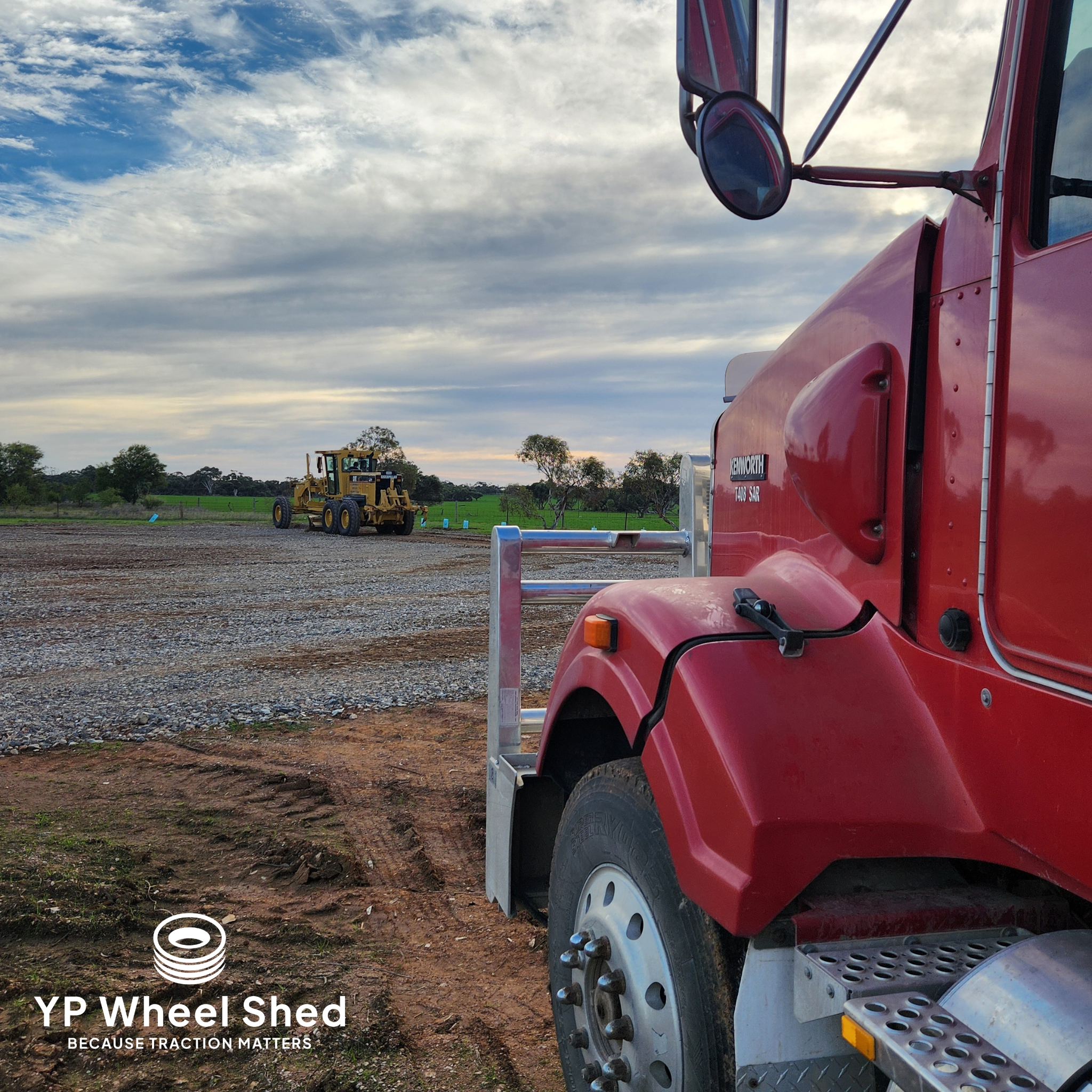 A red Kenworth prime mover parked on a gravel pad at YP Wheel Shed, with a grader working in the paddock behind it