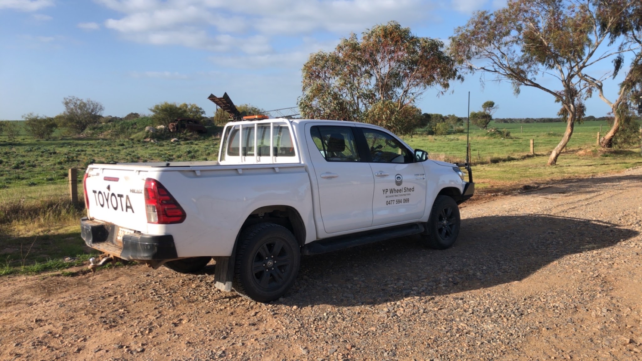 The signwritten YP Wheel Shed Hilux on a gravel track beside a green crop